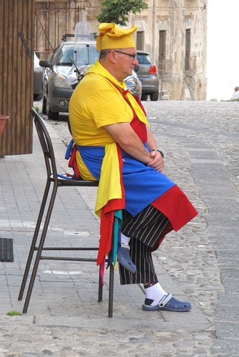 Immagine con giallo, strada, sedia, uomo, rosso, auto, blu, cuoco, cappello, grembiule, seduto, sgabello, colori, ciabatte, righe, occhiali, zoccoli, azzurro, multicolore