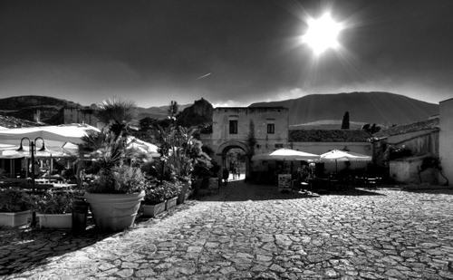 Immagine con sole, vasi, piazza, bianconero, piante, controluce, ombrelloni, arco, cielo, nero, finestre, porta, raggi, scorcio, case