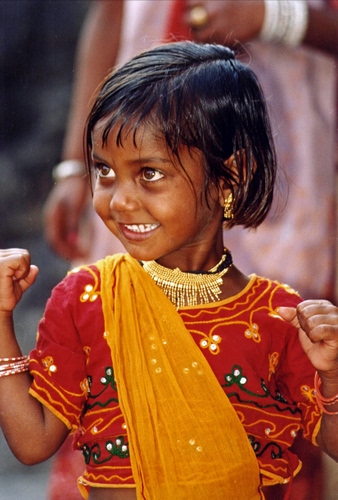 Immagine con bambina, sorriso, occhi, rosso, giallo, collana, sguardo, capelli, vestito, ritratto, mani, arancione, velo, viso, pugni