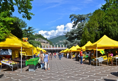 Immagine con mercato, giallo, alberi, nuvole, cielo, verde, tende, persone, piazza, bancarelle, azzurro, montagne, cassette, verdura, frutta, stand, ombrelloni