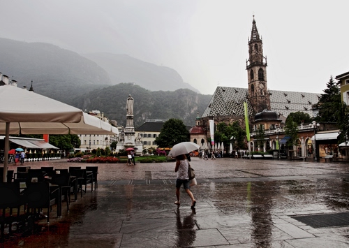 Immagine con campanile, ombrello, piazza, pioggia, chiesa, ombrelloni, sedie, monumento, montagne, passante, case, acqua, riflesso, statua, maltempo, ombrellone, persone